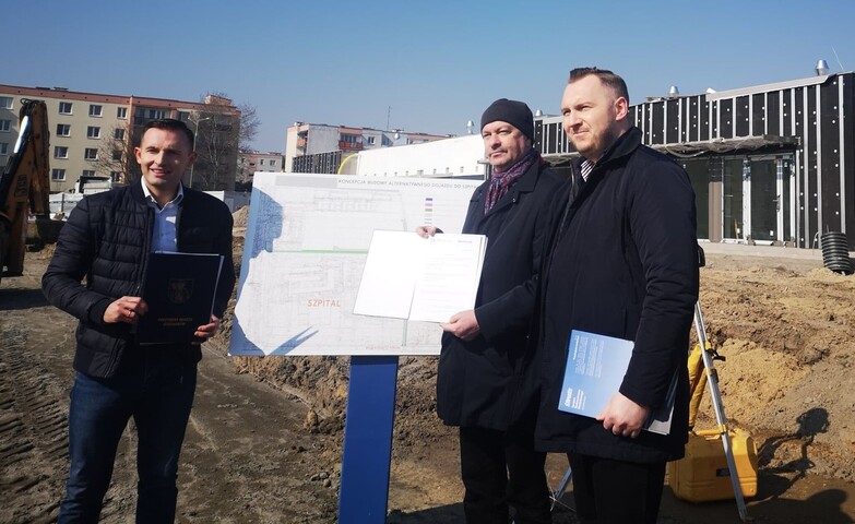 Trzech mężczyzn stoi na placu budowy, przy rozłożonym planie przebiegu drogi. Prezentują związane z nią dokumenty.