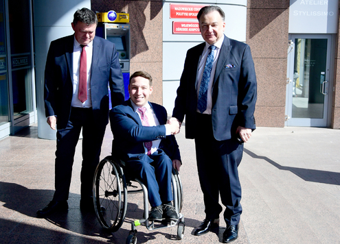Trzech mężczyzn w garniturach stoi obok siebie i pozuje do zdjęcia. Mężczyzna w środku siedzi na wózku inwalidzkim.