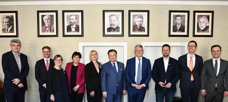 Marszałek, komisarz UE i prezydent Warszawy oraz pozostali uczestnicy spotkania stoją obok siebie i pozują do zdjęcia. Nad nimi wiszą portrety ważnych Europejczyków