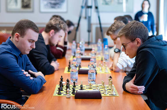 Widok na podłużny stół z czterema stanowiskami do gry w szachy. Na stole widać cztery plansze z pionkami, nad którymi pochylają się gracze
