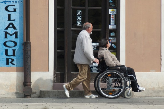 Mężczyzna prowadzi wózek inwalidzki, w którym siedzi kobieta. Idą chodnikiem obok schodów i drzwi