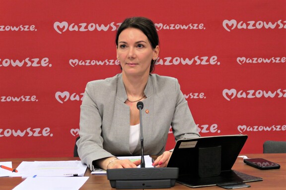 Radna siedzi za stołem obrad przed tabletem, za nią ścianka z logo samorządu Mazowsza
