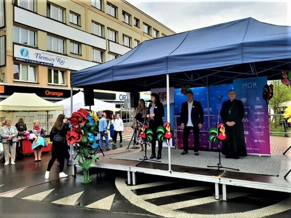 Scena na placu miejskim. Na scenie stoi Janina Ewa Orzełowska, która trzyma w ręku mikrofon. Za nią stoi mężczyzna oraz ksiądz. Przed sceną widać dziewczynę, która robi zdjęcia