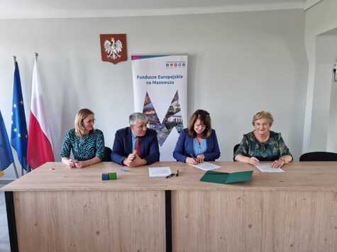 Cztery osoby, trzy kobiety i mężczyzna siedzą przy stole, dwie kobiety podpisują dokumenty