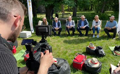 Grupa mężczyzn w słomkowych kapeluszach siedzi na leżakach na trawie. Filmuje ich operator kamery.