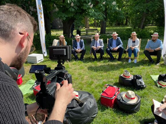 Grupa mężczyzn w słomkowych kapeluszach siedzi na leżakach na trawie. Filmuje ich operator kamery.