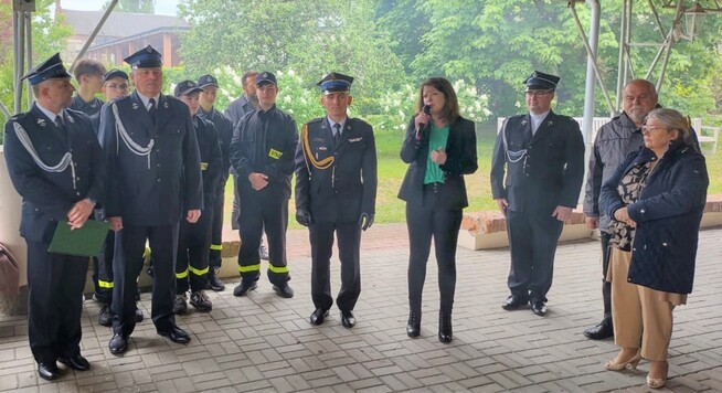 kobieta z mikrofonem w ręku przemawia na placu, wokół niej stoi grupa strażaków w odświętnych mundurach