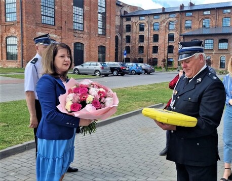 Janina Ewa Orzełowska stoi z bukietem kwiatów przed strażakiem, który trzyma w rękach pudełko z sygnalizatorami.