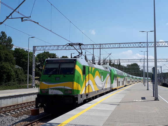 Zielono-żółty pociąg stoi na peronie. W tle błękitne niebo.