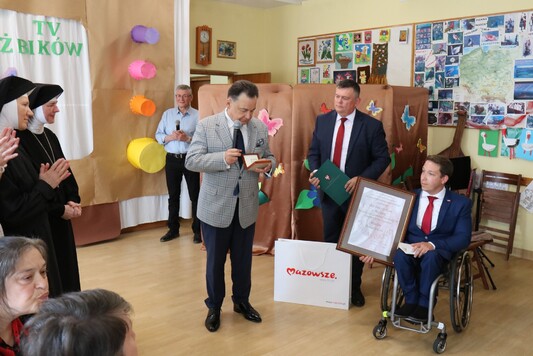 Marszałek stoi i trzyma pudełko z medalem, obok iego jest dyrektor Kornatowski, który trzyma list, a obok niego na wózku inwalidzkim siedzi Artur Świercz, który trzyma dyplom Pro Masovia