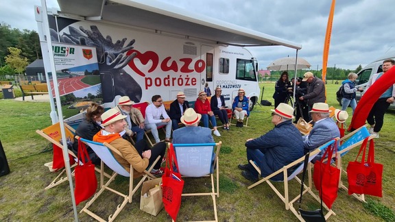 Grupa osób w słomkowych kapeluszach siedzi na leżakach, rozstawionych na polanie przy białym kamperze z logo Mazowsza