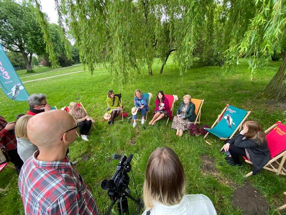 Janina Ewa Orzełowska, Elżbieta Lanc, dyrektor Stelmańska i rzeczniczka UMWM oraz dwie dziewczyny siedzą na leżakach ustawionych w parku pod wierzbą. Przed nimi widać mikrofony dziennikarzy, którym udzielają wywiadu