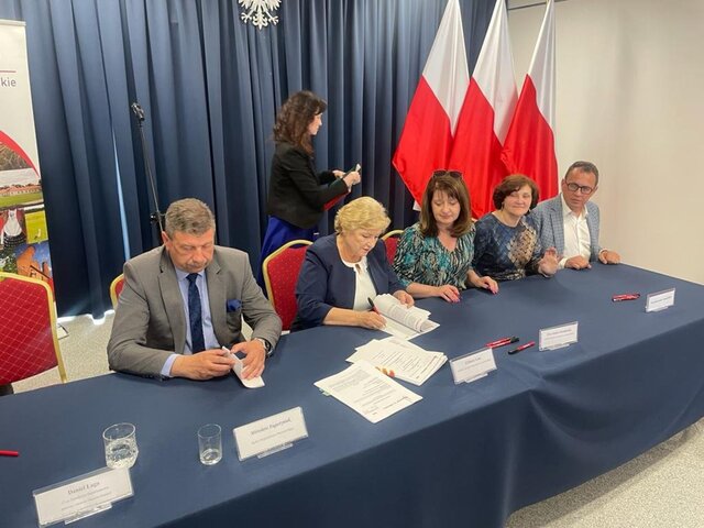 5 osób siedzi przy stole i podpisuje umowy na tle niebieskiego tła z flagami Polski