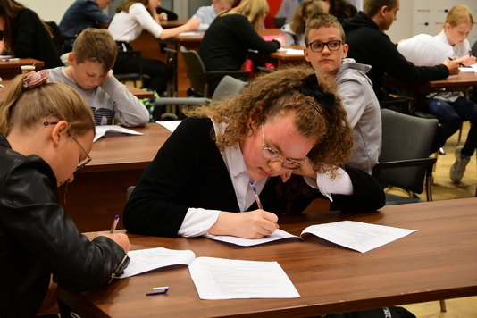 Uczennica siedzi przy stoliku nachylona nad kartką, na której trzyma prawą dłoń z długopiem. Obok niej widać innych uczniów biorących udział w teście