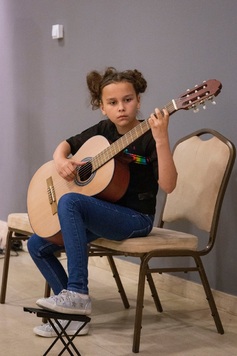 Dziewczynka siedzi na krześle, lewą stopę trzyma na podnóżku. W rękach trzyma gitarę klasyczną.