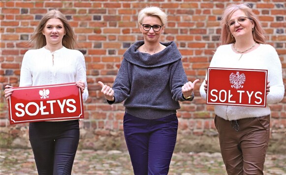 Trzy kobiety, dwie trzymają tabliczkę z napisem 
