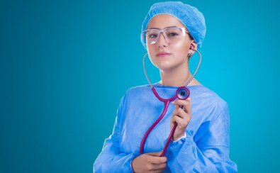 Młoda lekarka w fartuchu, czepku, okularach ochronnych i stetoskopem w ręku patrzy się przed siebie