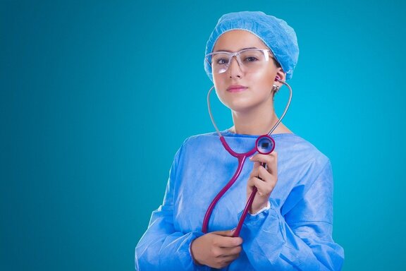Młoda lekarka w fartuchu, czepku, okularach ochronnych i stetoskopem w ręku patrzy się przed siebie