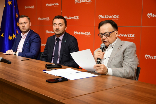 Trzech mężczyzn siedzi przy stole, jeden z nich mówi do mikrofonu. W tle logotypy Mazowsza.