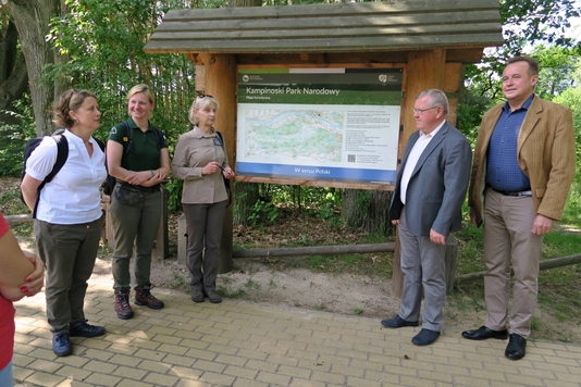 Radni i przedstawiciele Kampinoskiego Parku Krajobrazowego stoją przy tablicy informacyjnej.
