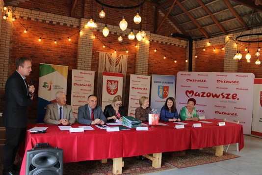 Beneficjenci i przedstawiciele samorządu województwa mazowieckiego siedzą za stołem na tle ścianki z logo Mazowsza i podpisuja umowy