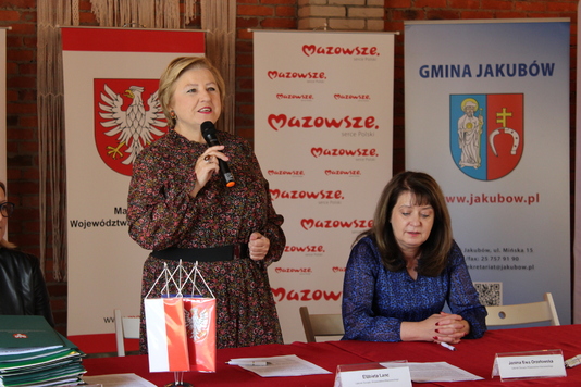 Elżbieta lanc soti za stołem przemawiając do mikrofony, Janina Ewa Orzełowska siedzi obok przy tym samym stole