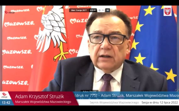 Stopklatka z transmisji internetowej. Marszałek mówi siedząc za biurkiem, za nim flagi Polski, Mazowsza i Unii Europejskiej oraz ścianka z logotypem samorządu Mazowsza.
