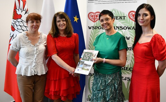 Janina Ewa Orzełowska pozuje do zdjęcia z trzema kobietami. Za nim są flagi Polski i UE oraz baner KSOW