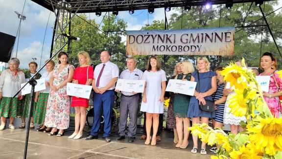 Na scenie stoi Janina Ewa Orzełowska oraz sześciu przedstawicieli miasta Mokobody. Pozują do zdjęcia, stojąc obok siebie. Za nimi jest baner Dożynki gminne mokobody