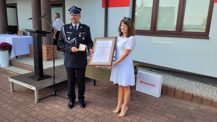 Janina Ewa Orzełowska stoi obok strażaka w oficjalnym strażackim mundurze. Oboje trzymają dyplom Pro Masovia