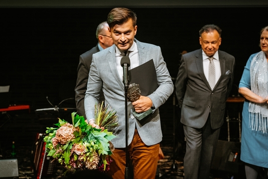 Jakub Małecki stoi przy mikrofonie. W prawym ręku trzyma kwiaty, w lewym ręku dyplom i statuetkę. W tle widać marszałka Adama Struzika.