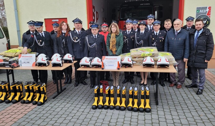 Orzełowska pozuje do zdjęcia ze strażakami z OSP Mlęcin i Dobre. Przed nimi na stole rozłożone są komplety umundurowania bojowego