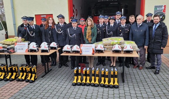 Orzełowska pozuje do zdjęcia ze strażakami z OSP Mlęcin i Dobre. Przed nimi na stole rozłożone są komplety umundurowania bojowego