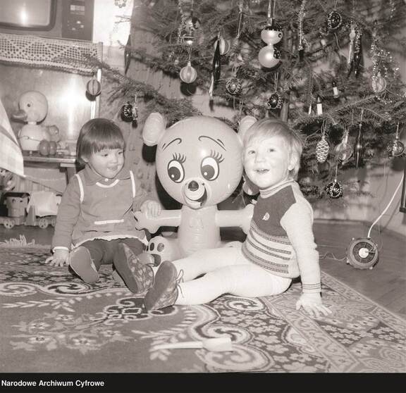 dwójka dzieci w wieku przedszkolny siedzi na dywanie. Przytulają dmuchaną zabawkę w kształcie misia. W tle udekorowana choinka