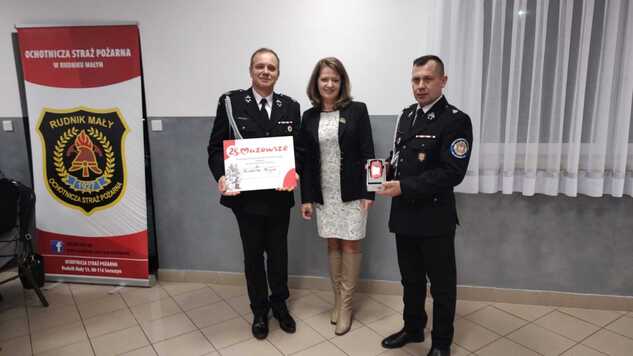 Członek zarządu Janina Ewa Orzełowska z dwoma strażakami. Jeden z nich trzyma tablicę informującą o wsparciu