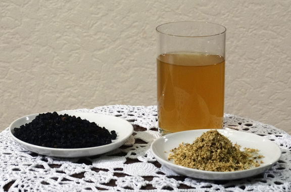 Szklanka z herbatą, a obok niej na dwóch talerzykach pokruszone kwiaty i owoce czarnego bzu