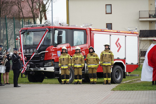 Wóz strażacki, a obok niego czterech strażaków w uniformach