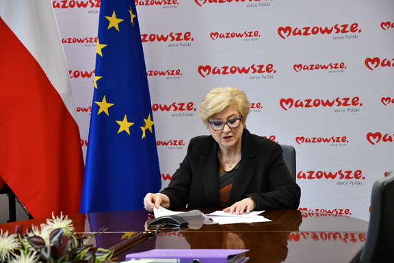 Elżbieta Lanc, członek zarządu województwa, siedzi za stołem. Przed sobą ma dokumenty, które przegląda. W tle widać flagi UE, Polski i Mazowsza oraz baner z napisem Mazowsze